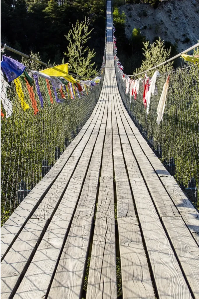Das Bild zeigt eine Hängebrücke aus Holz. An den Seiten hängen bunte Tücher.